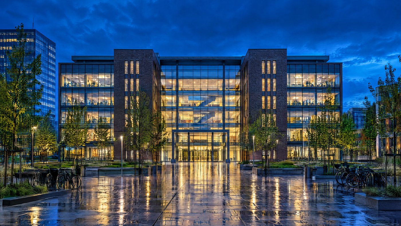 The VU Amsterdam campus in the Zuidas at blue hour, lit windows reflected in a wet plaza with bicycles parked out front.
